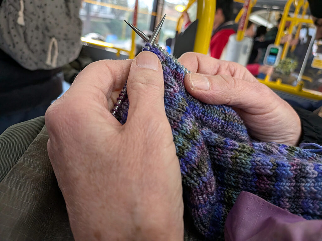 Close-up of a person's hands knitting on a bus with steel circular needles and variegated yarn in shades of purple, blue, and mossy green.