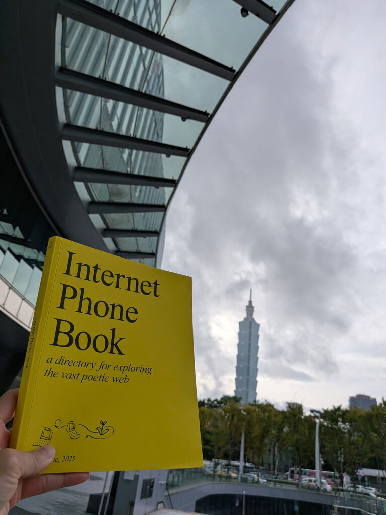 A hand holds a bright yellow copy of the Internet Phone Book against a grey, cloudy sky, with the tiered architecture of the Taipei 101 tower in the background.
