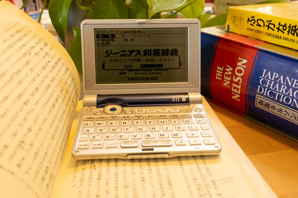 A vintage silver Seiko SR-MK4100 electronic dictionary open on a wooden desk. It sits on top of an open Japanese book, flanked by a thick red 'New Nelson' Japanese-English Character Dictionary and a yellow 'Furigana English-Japanese' dictionary. The electronic screen shows the 'Genius' dictionary interface in monochrome.